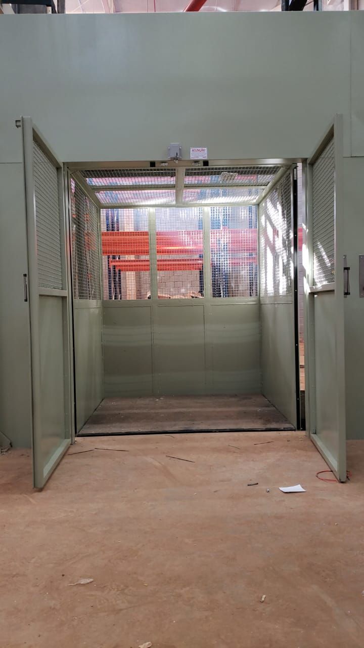 Elevador de carga com portas metálicas verde-claras, mostrando a plataforma interna contra um fundo vermelho.