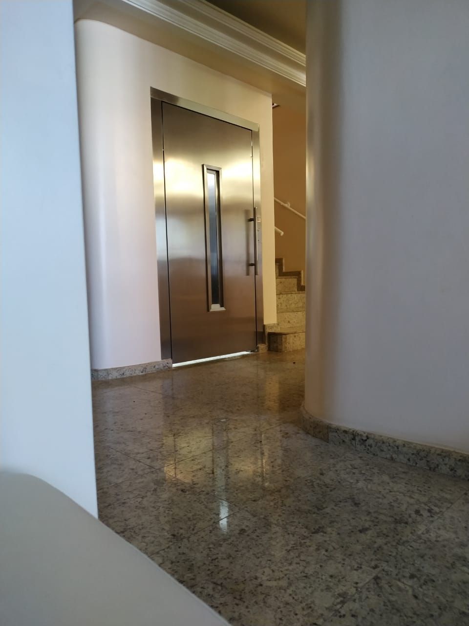 Porta de elevador em aço inoxidável em um corredor com piso de granito polido e paredes curvas brancas.