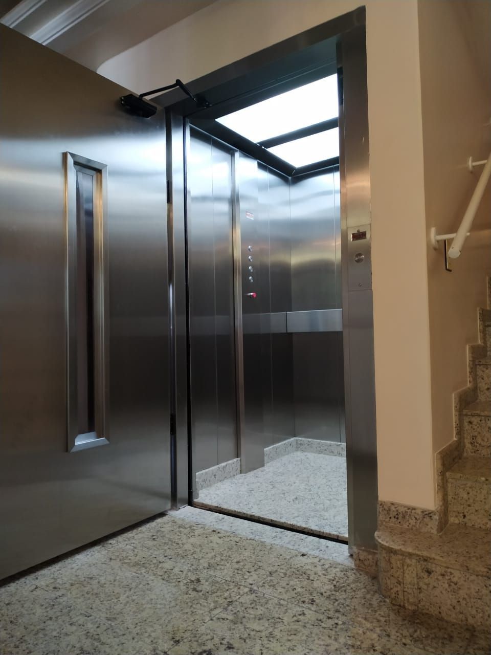 Portas de elevador de aço inoxidável abertas em um corredor com piso de ladrilho de pedra ao lado de uma escada.