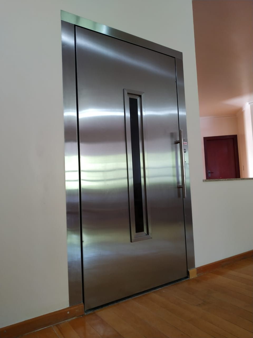 Porta de elevador em aço inoxidável com um painel de vidro vertical, instalada em uma sala simples com piso de madeira.