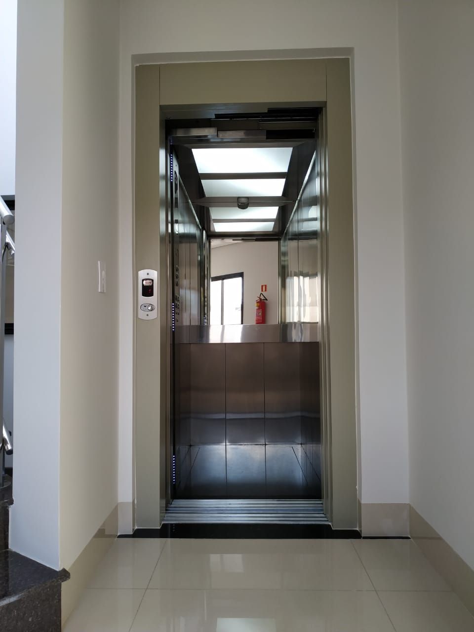 Porta de elevador aberta em um corredor com paredes brancas e piso de azulejos claros.