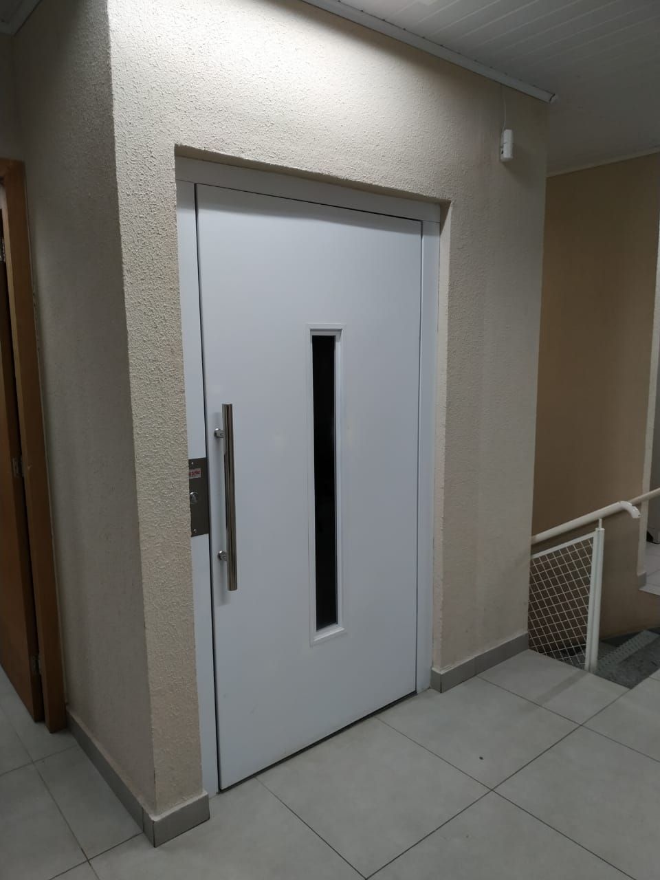 Porta de elevador branca, com painel de vidro vertical e puxador de metal, localizada em um corredor interno de tons neutros.
