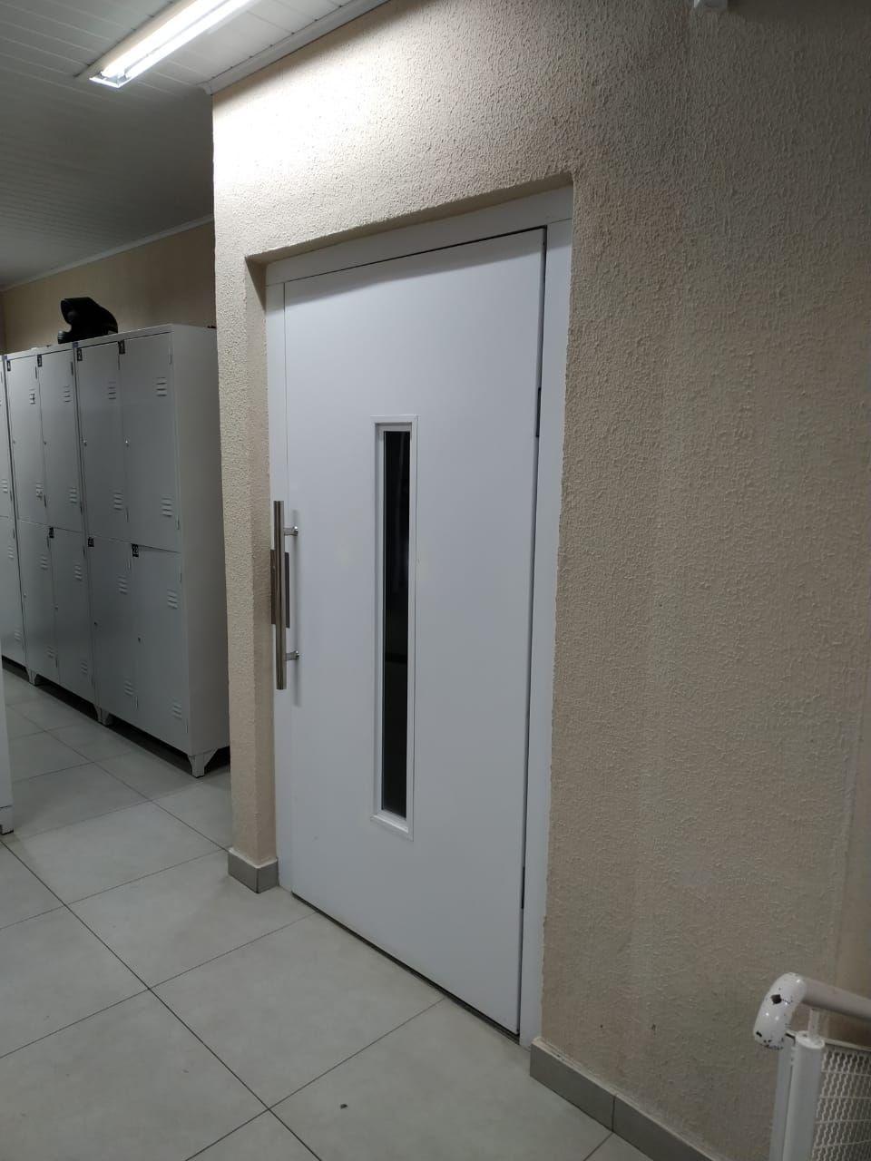 Porta branca com um painel de vidro vertical e uma maçaneta prateada, situada em um corredor ao lado de armários de metal.