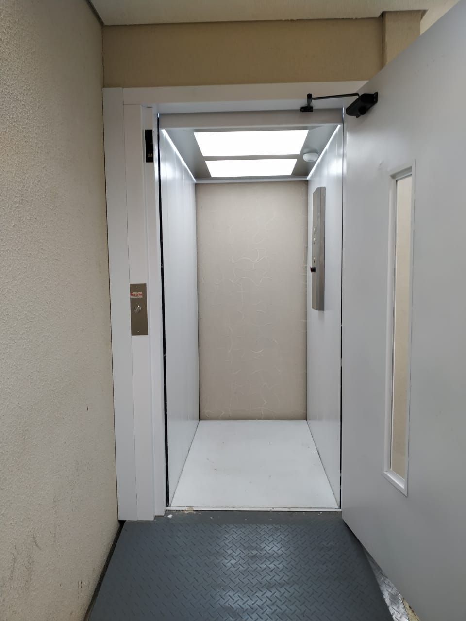 Porta de elevador aberta revelando um interior pequeno, bem iluminado e vazio, com paredes brancas e piso texturizado cinza.
