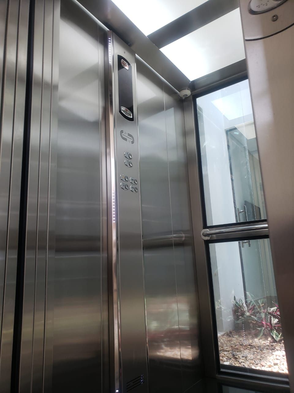 Interior de um elevador de aço inoxidável com painel de seleção de andares e parede de vidro com vista para a vegetação.