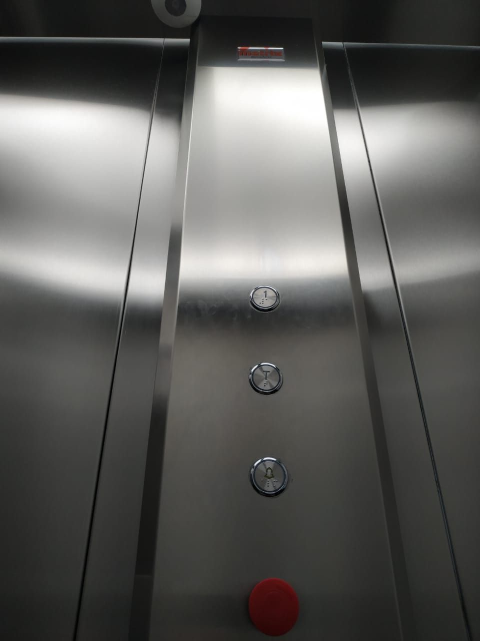 Painel de controle de elevador em aço inoxidável com três botões circulares e um botão vermelho de emergência abaixo.