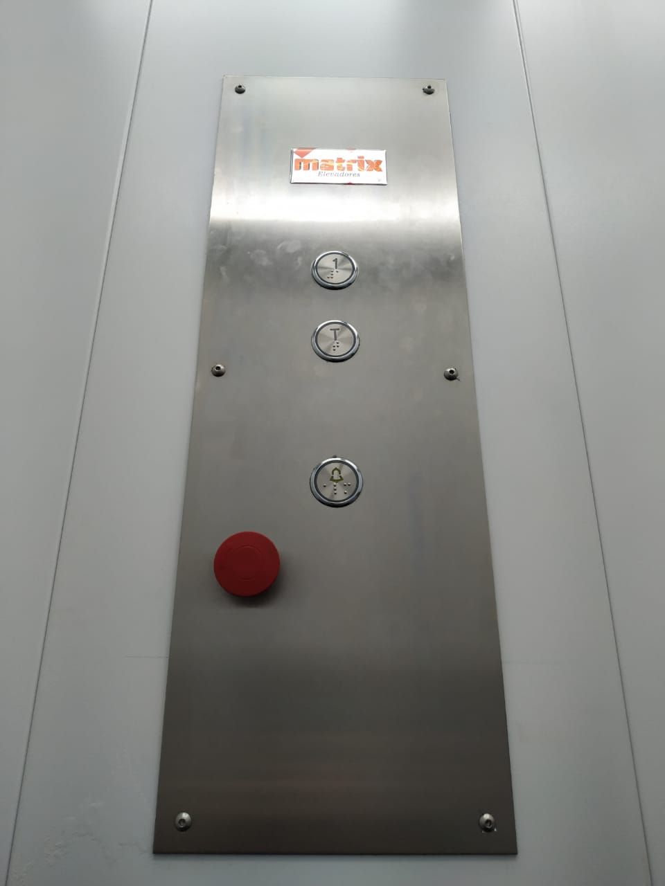Painel vertical de controle de elevador em metal numa parede, com três botões circulares e um botão de emergência vermelho.
