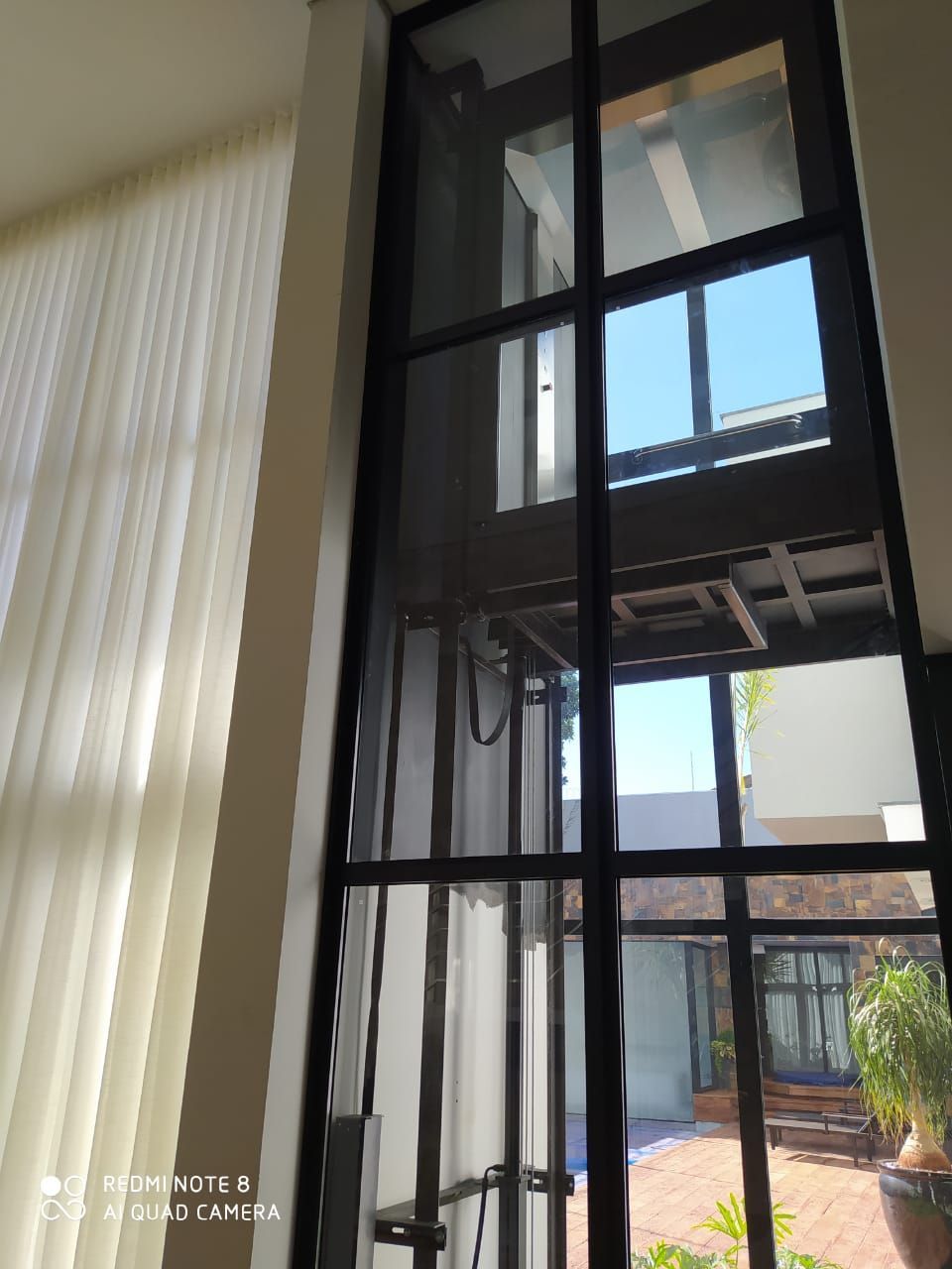 Elevador residencial com estrutura metálica preta e painéis de vidro, com vista para um pátio ensolarado e com plantas.