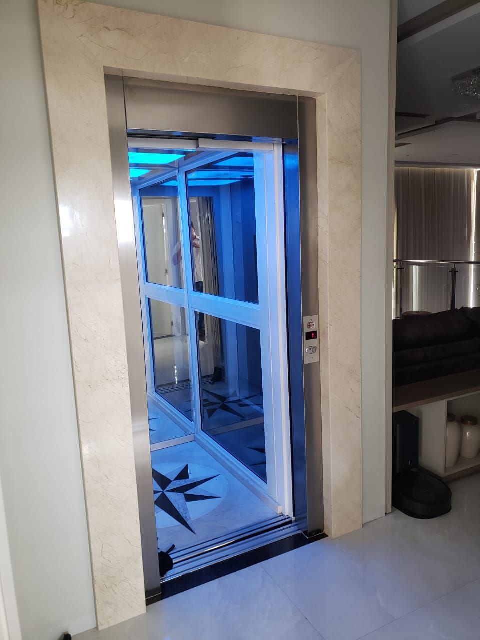 Elevador residencial com portas de vidro e iluminação interna azul, emoldurado por uma ampla borda de mármore.