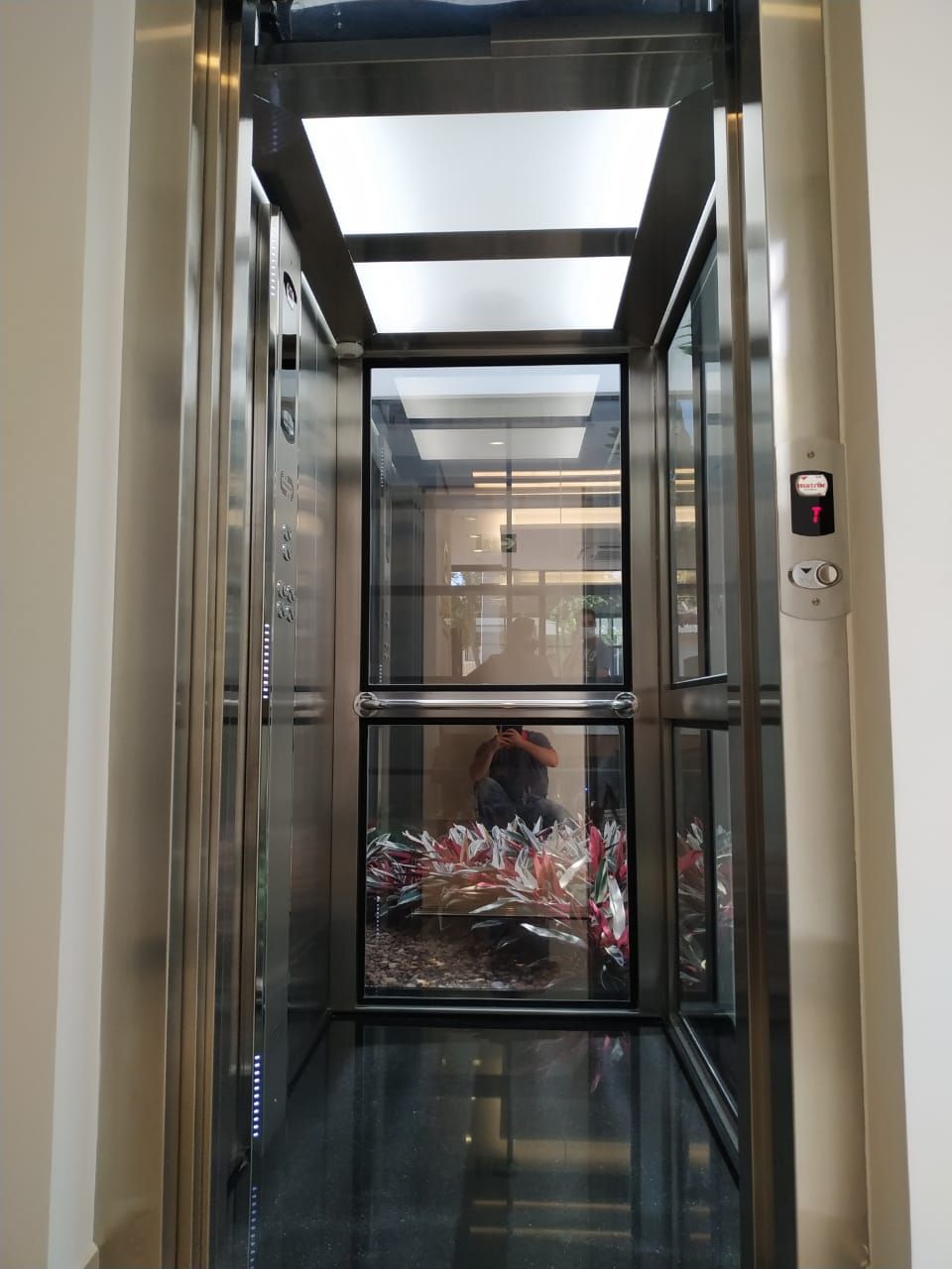 Elevador moderno com paredes de vidro e estrutura metálica, que exibe o reflexo de uma pessoa em seu interior.