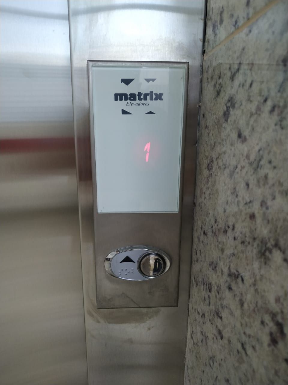 Painel de chamada de elevador em metal escovado com visor digital exibindo o número um e um botão de seta para cima.