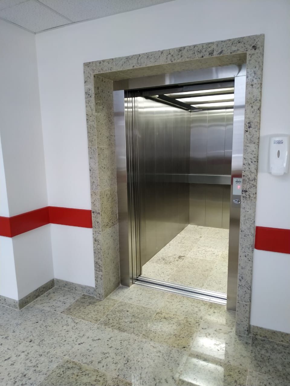 Porta de elevador aberta, em uma parede branca com detalhes em vermelho, piso e moldura da porta de pedra salpicada.