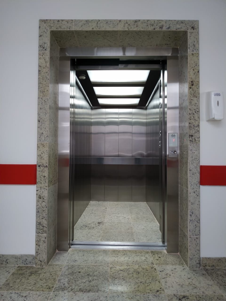 Porta de elevador aberta em aço inoxidável, emoldurada por pedra cinza-clara com uma faixa horizontal vermelha na parede.
