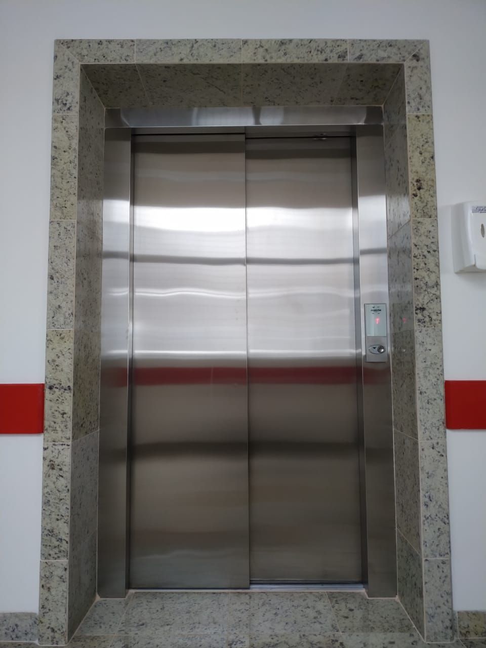 Elevador fechado com portas de aço inoxidável emolduradas por granito, encostado a uma parede branca com uma faixa vermelha.