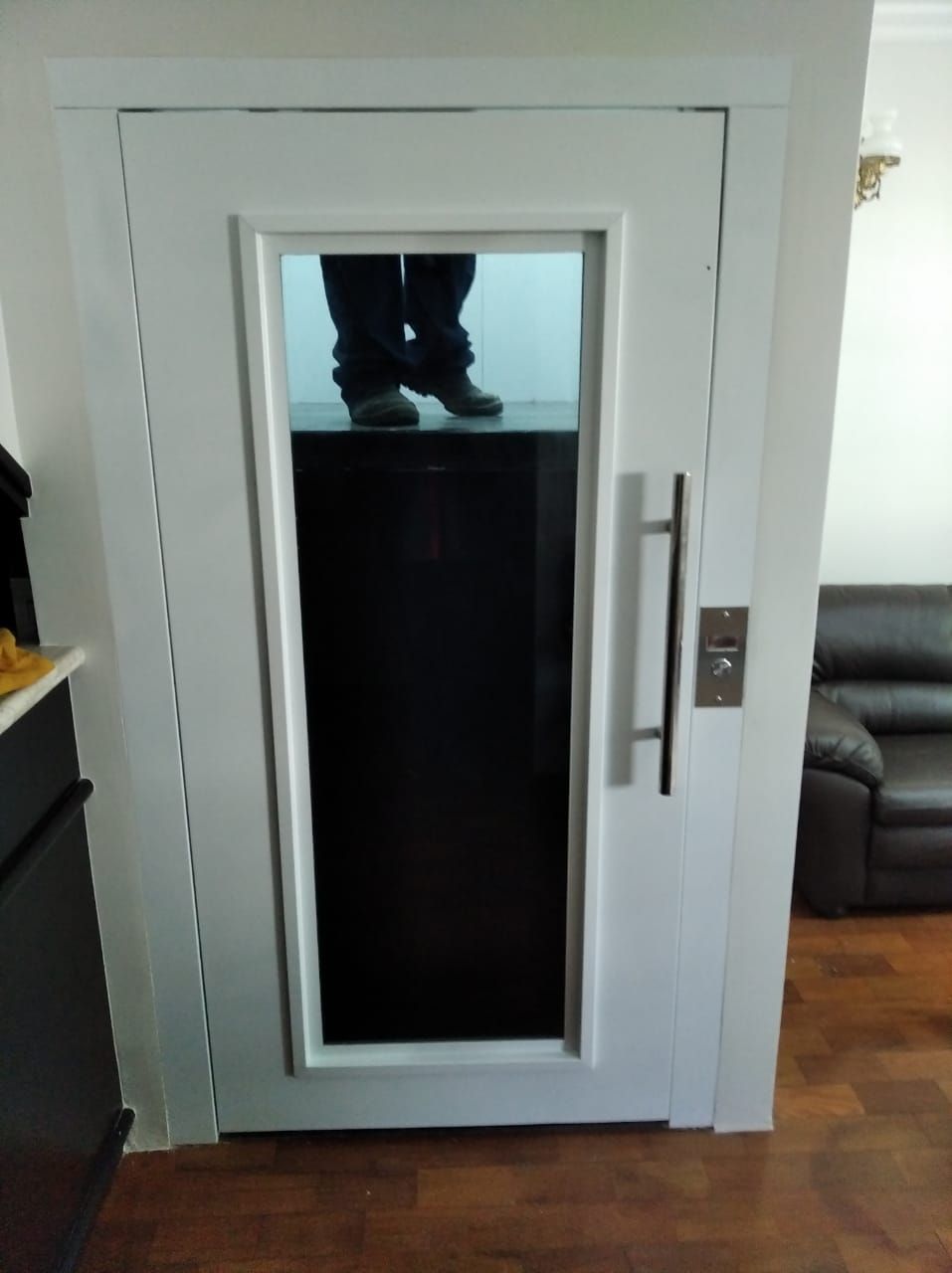 Porta de elevador residencial branca com janela de vidro vertical e uma longa alça de metal.