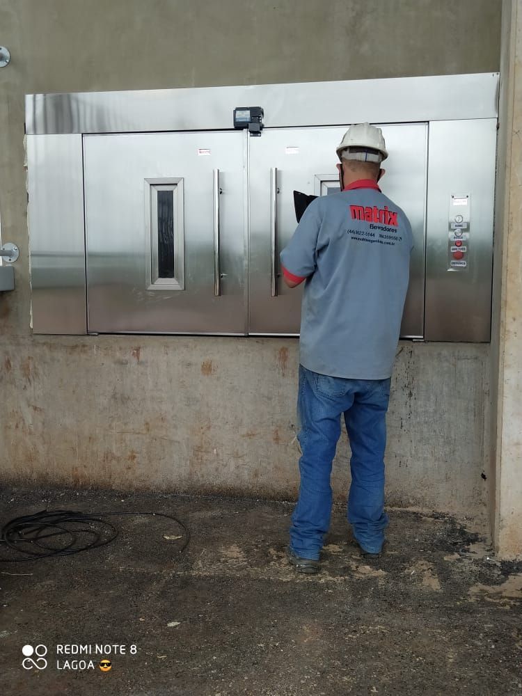 Pessoa usando capacete e camisa cinza inspeciona a instalação de uma porta dupla de aço inoxidável em uma parede inacabada.