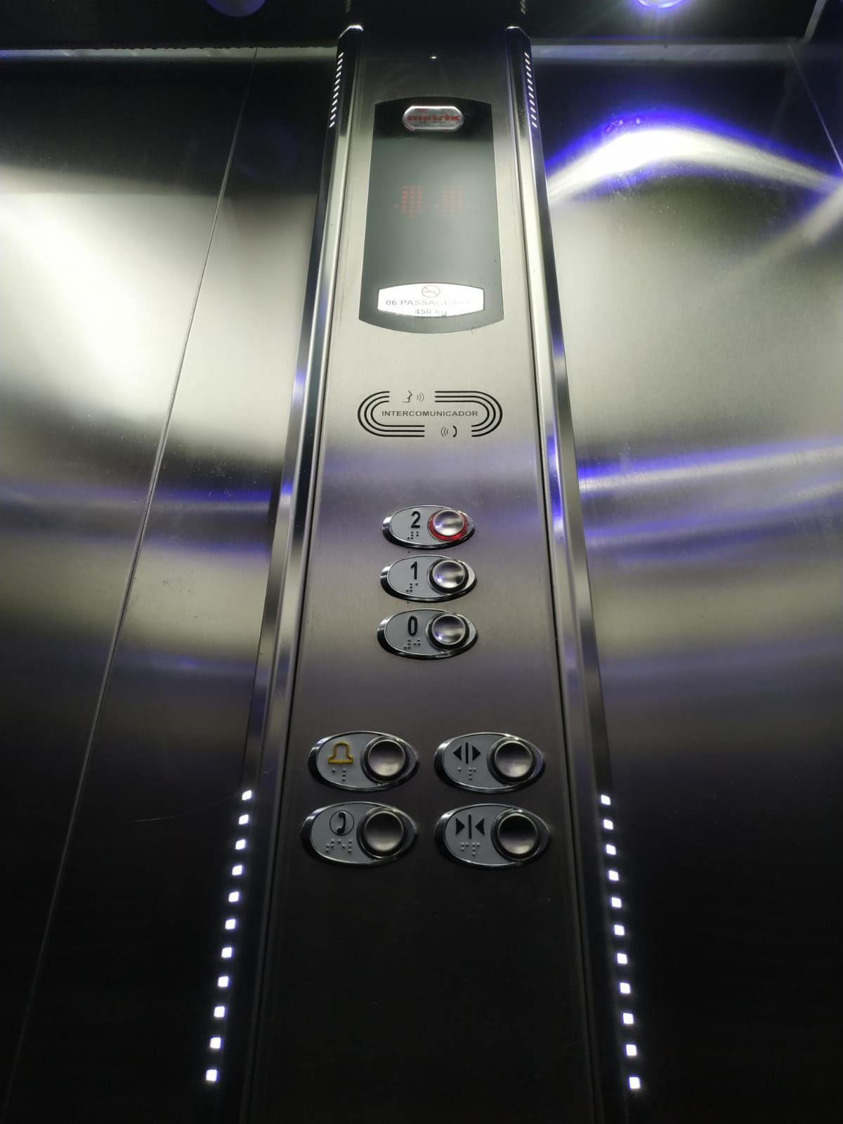 Painel de controle de elevador com três botões de andar, um botão para abrir/fechar e um botão de alarme de emergência.
