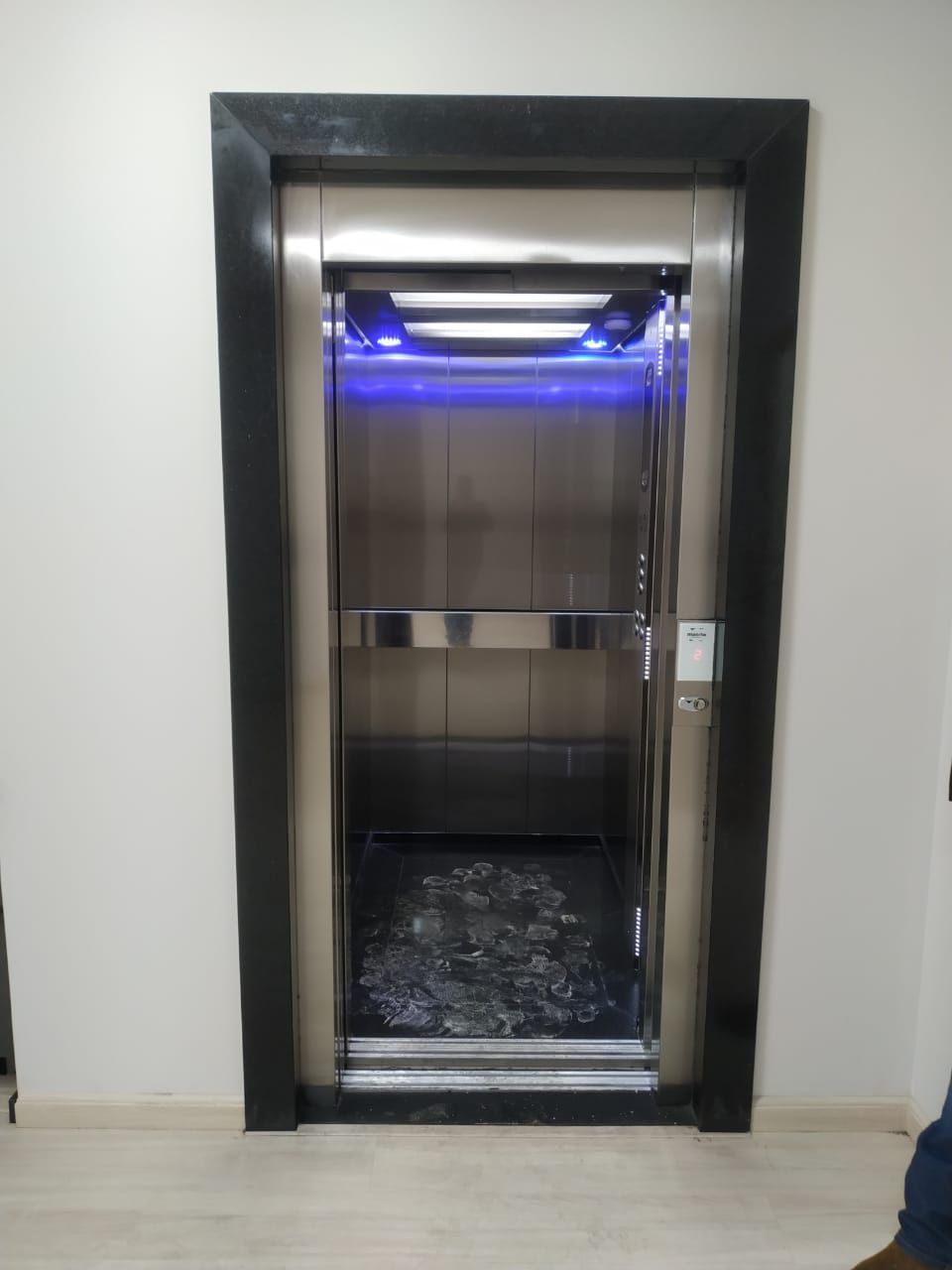 Elevador aberto com portas de aço inoxidável, iluminação interior moderna e piso escuro, situado num corredor branco.