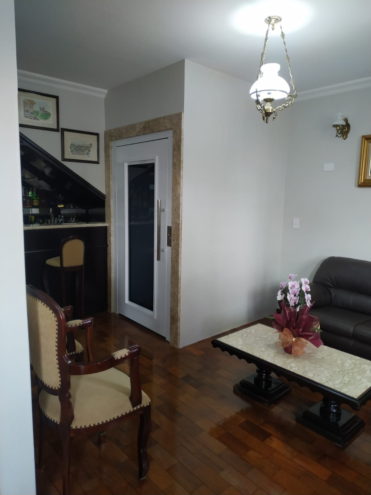 Sala de estar com piso de madeira, uma cadeira bege, um sofá pequeno, uma mesa de centro com flor e uma porta de elevador.