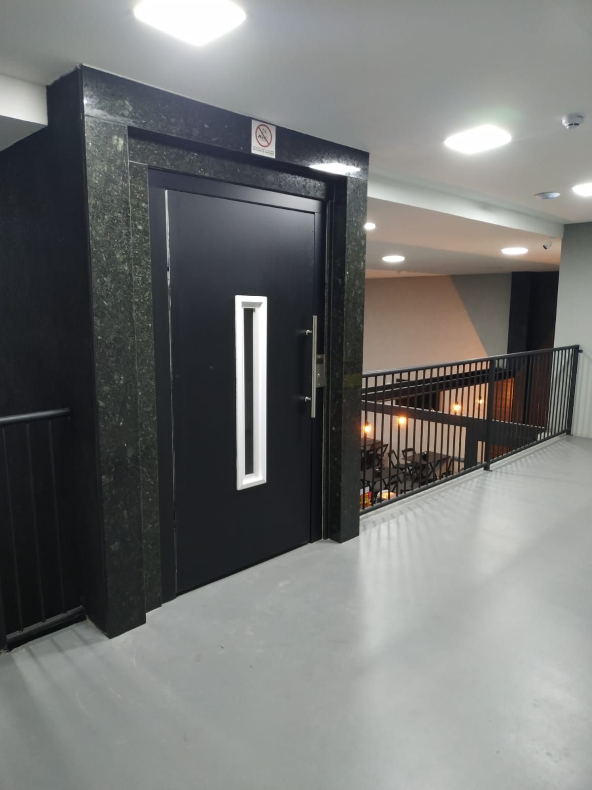 Porta de elevador preta com painel de vidro vertical, em um cubo de mármore, no corredor de um edifício ao lado do corrimão.