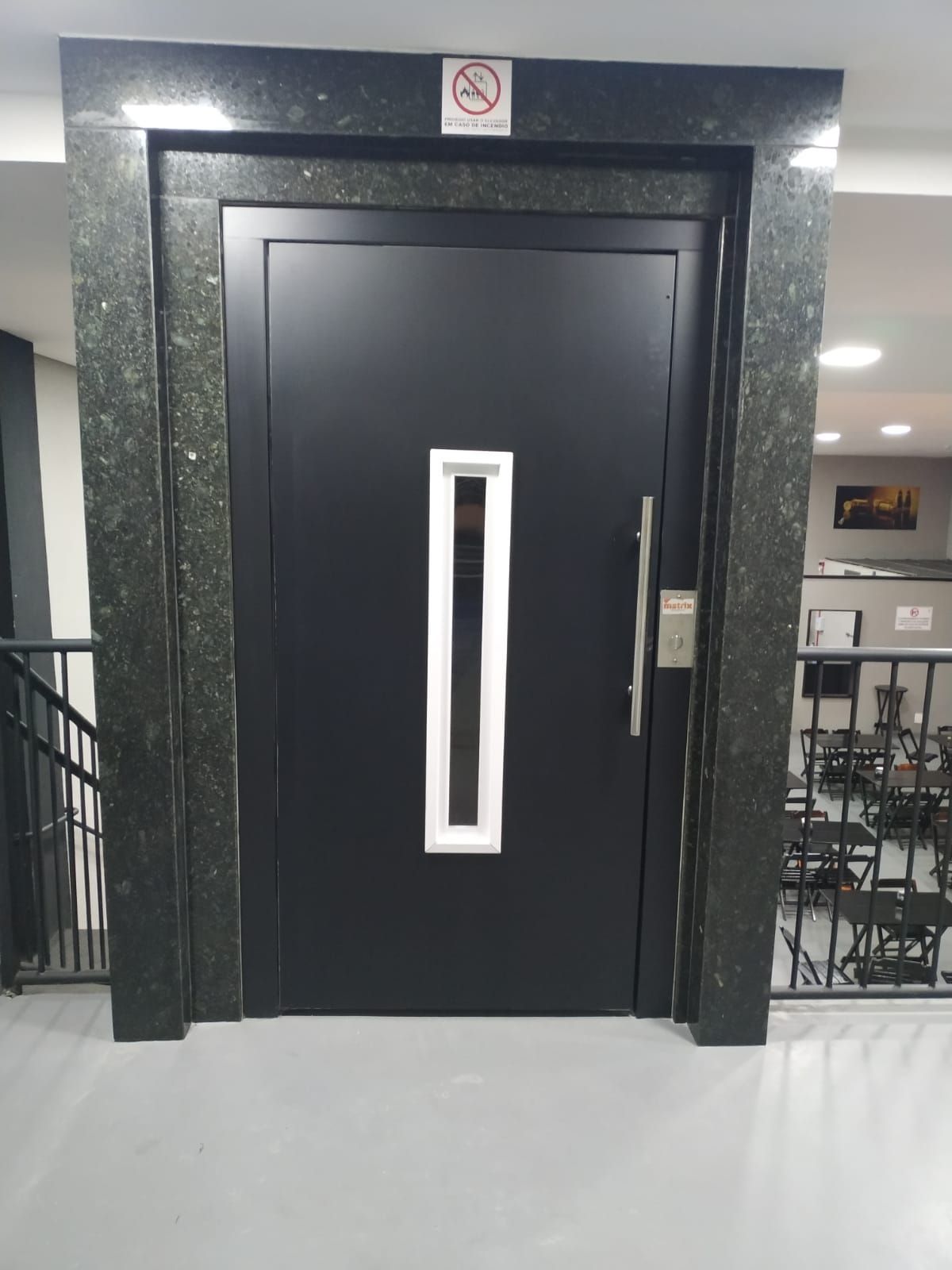 Porta de elevador preta com painel de vidro vertical, inserida numa moldura de mármore, num corredor público.