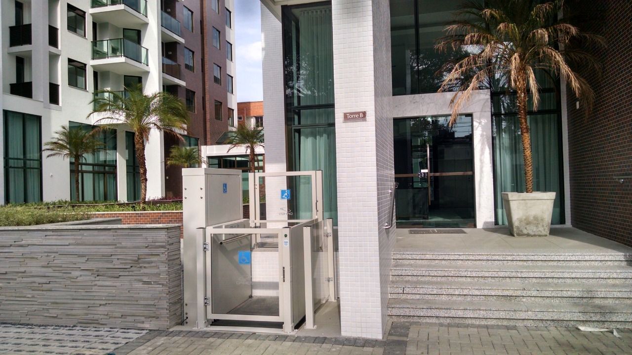 Plataforma elevatória vertical para cadeiras de rodas instalada ao lado da escadaria da entrada de um prédio moderno.