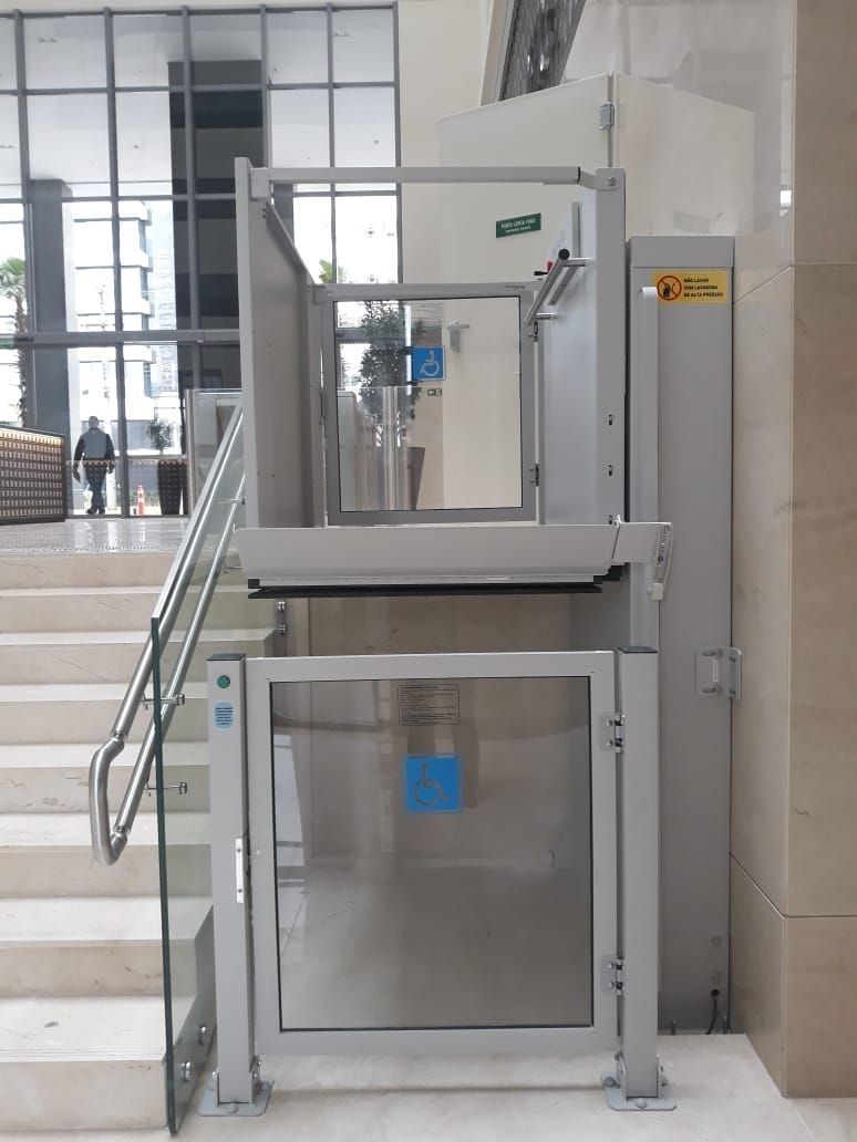 Plataforma elevatória cinza para cadeiras de rodas instalada ao lado de uma escada no saguão de um prédio bem iluminado.