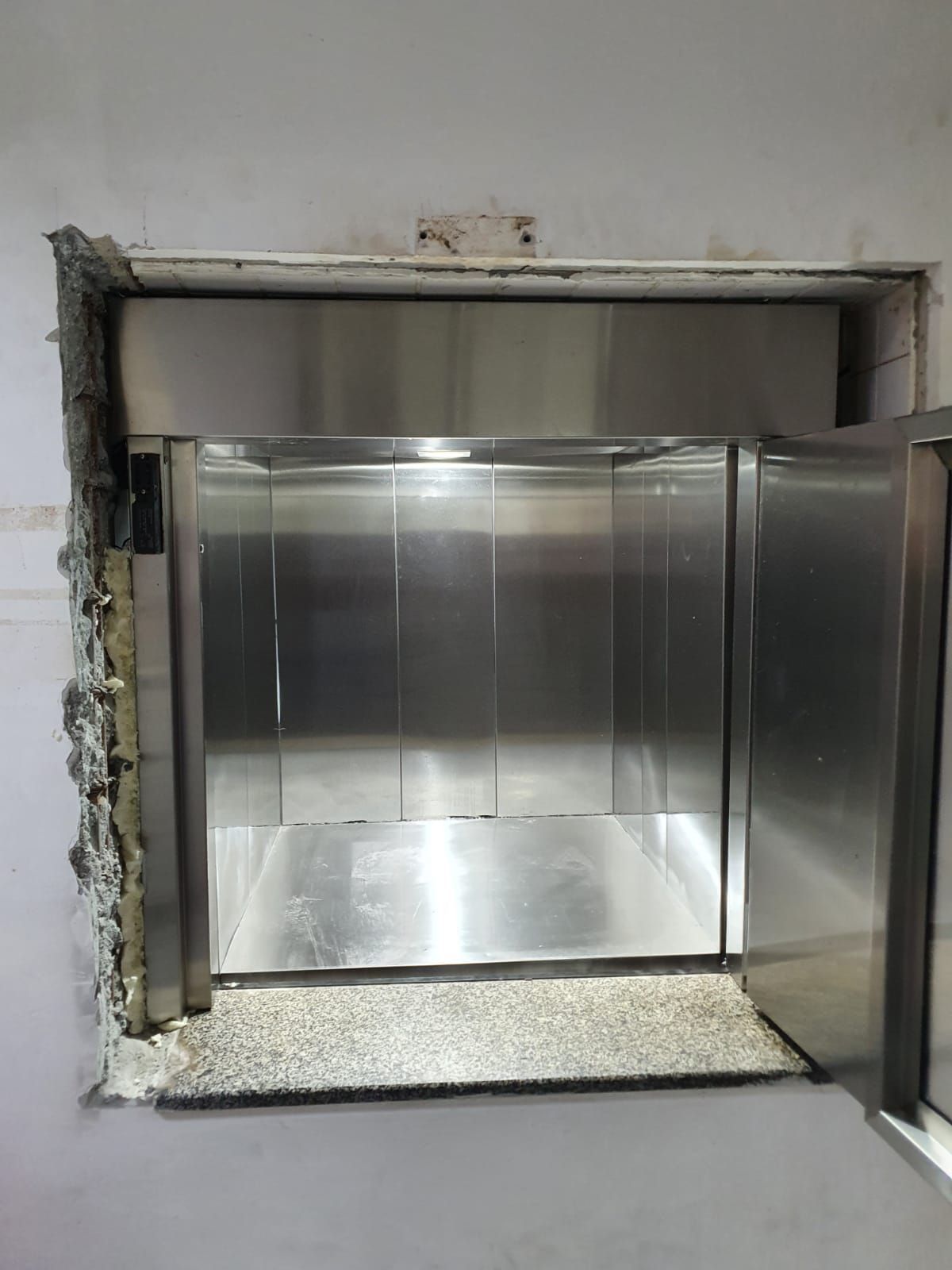 Elevador monta-cargas de aço inoxidável com porta aberta, instalado em parede parcialmente acabada.