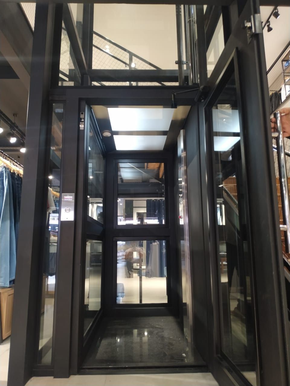 Elevador moderno com paredes de vidro e estrutura metálica preta, instalado dentro de uma loja de roupas.