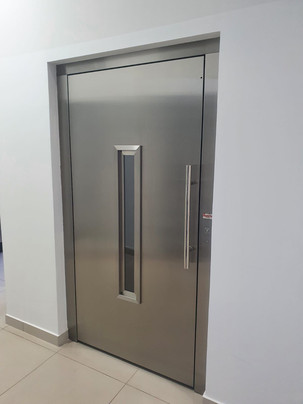Porta de elevador de aço inoxidável com uma janela vertical e uma alta maçaneta prateada, instalada em uma parede branca.