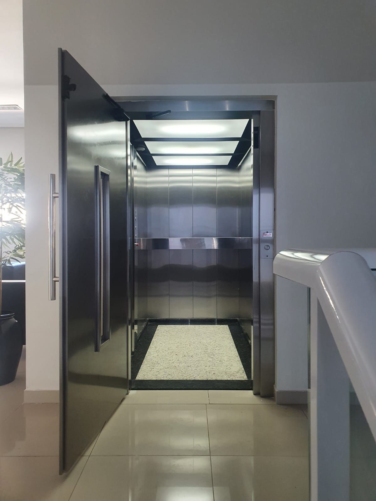 Elevador aberto com interior em aço inoxidável e piso de seixos brancos, visível da área do saguão adjacente.