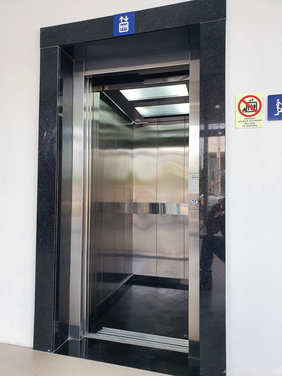 Elevador aberto com paredes de aço inoxidável, acabamento em mármore preto e sinalização acessível em uma parede branca.