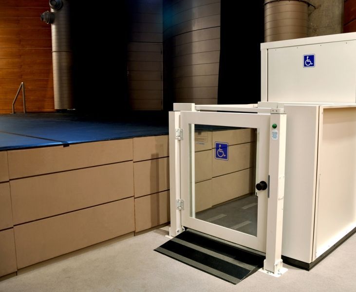 Elevador adaptado para cadeirante de rodas, posicionado ao lado de um palco de madeira elevado em ambiente interno.