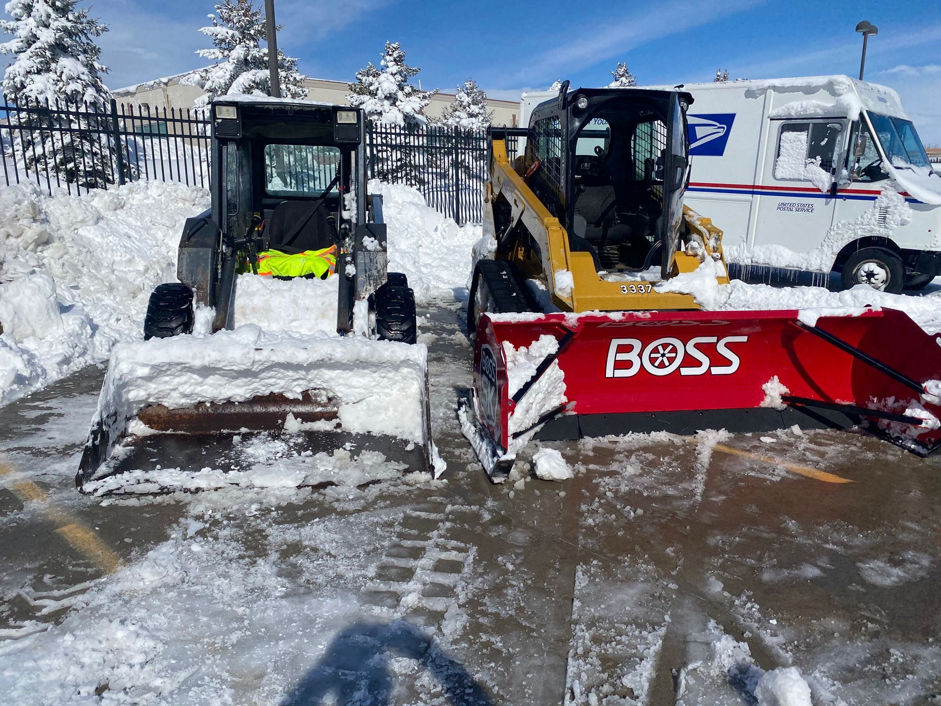 Two snowplows with red and yellow blades parked in a snowy lot, with a white van behind them.