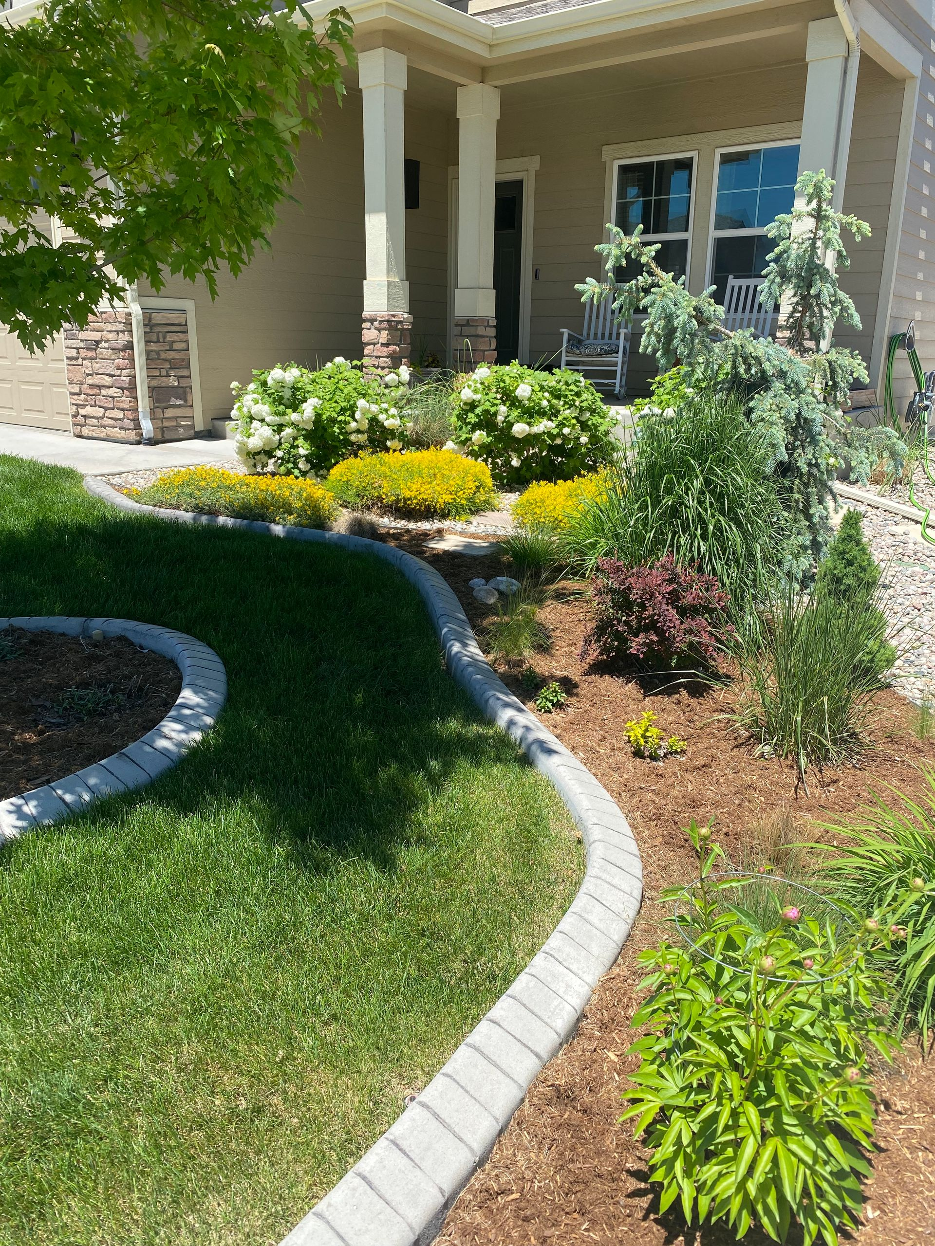 Front yard garden with curving stone border, green lawn, shrubs, and a house porch in sunlight