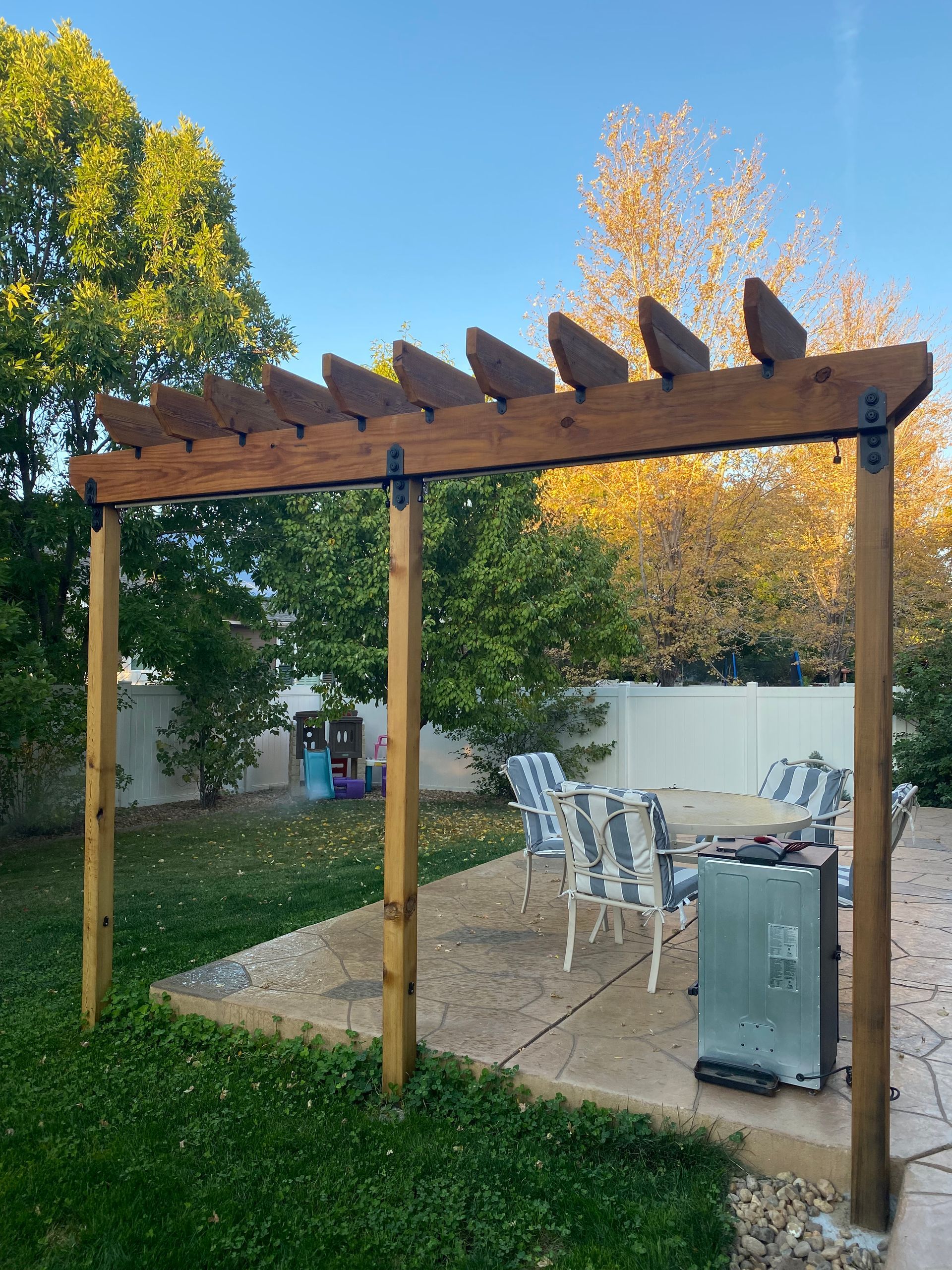 Wooden pergola in a backyard with patio chairs, a grill, and trees in autumn light