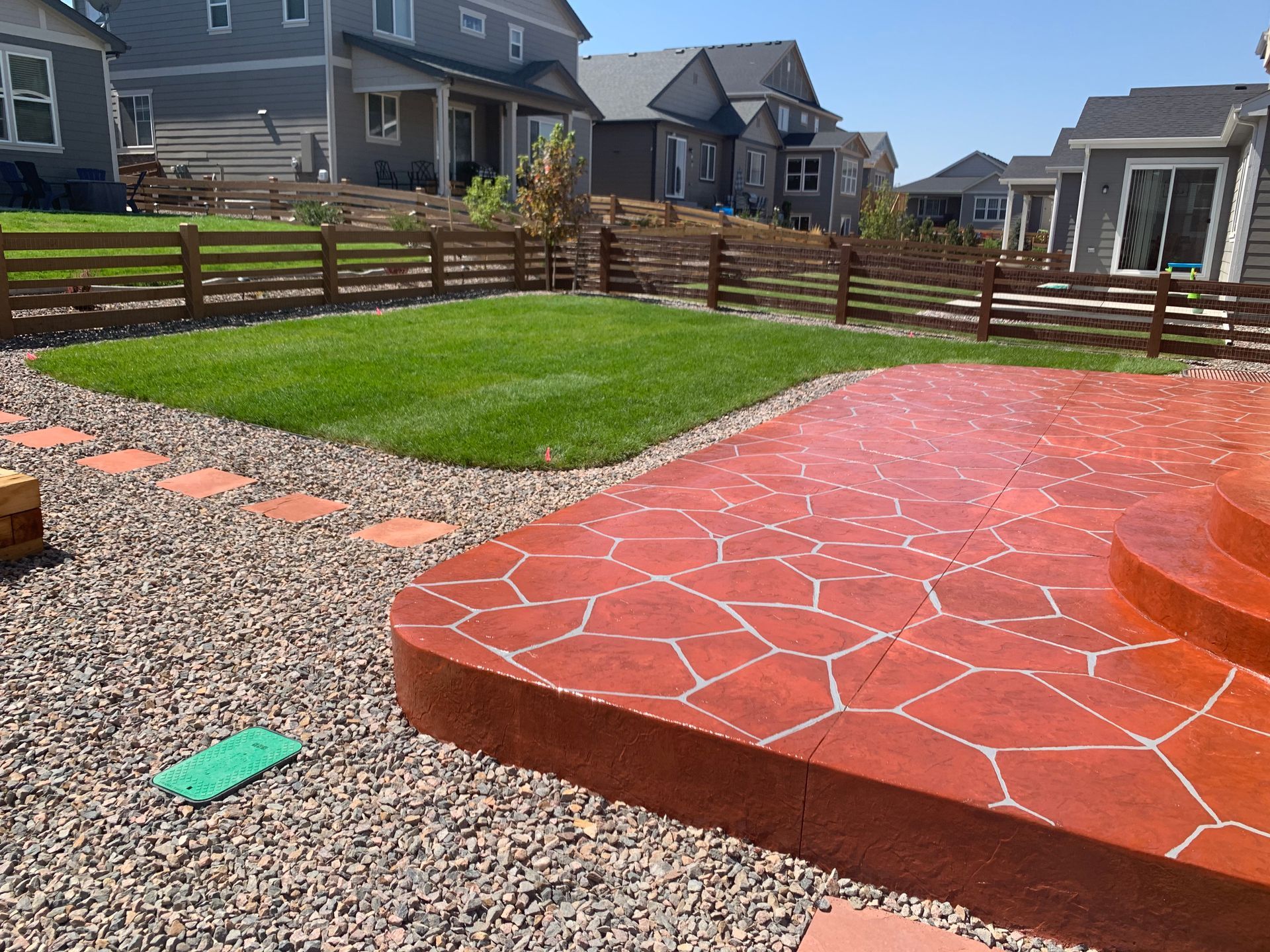 Backyard patio with red stamped concrete, gravel path, green lawn, and wooden fence in a suburban neighborhood