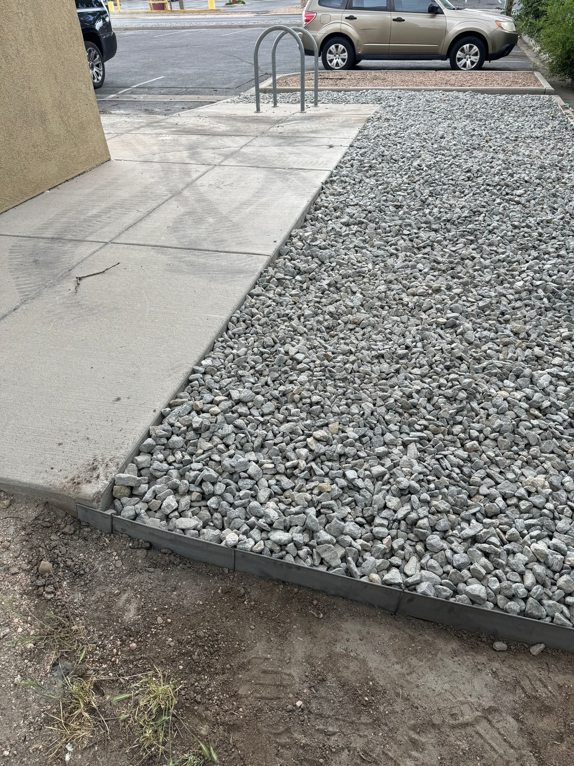 Concrete sidewalk beside a gravel driveway and parked car on a wet, overcast street