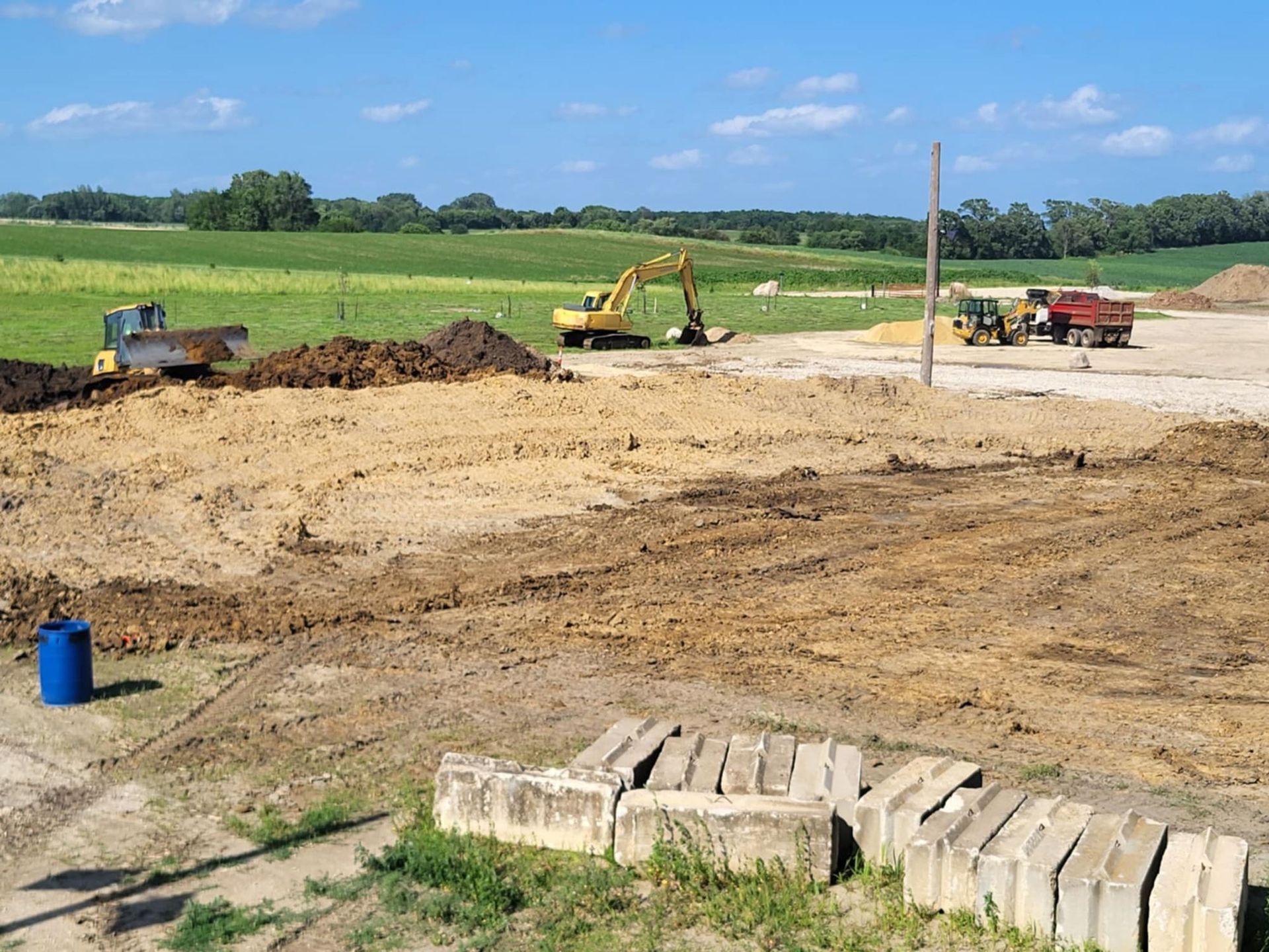 Construction site with excavator, dump truck, and dirt piles in a grassy field under blue sky