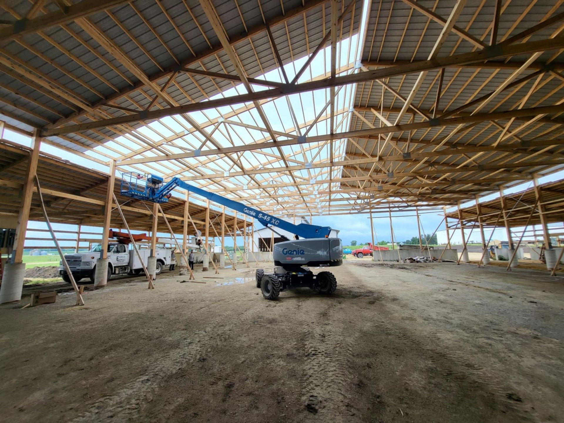 Inside a large open-sided barn, a blue cherry picker sits on a dirt floor with trucks along the wall.