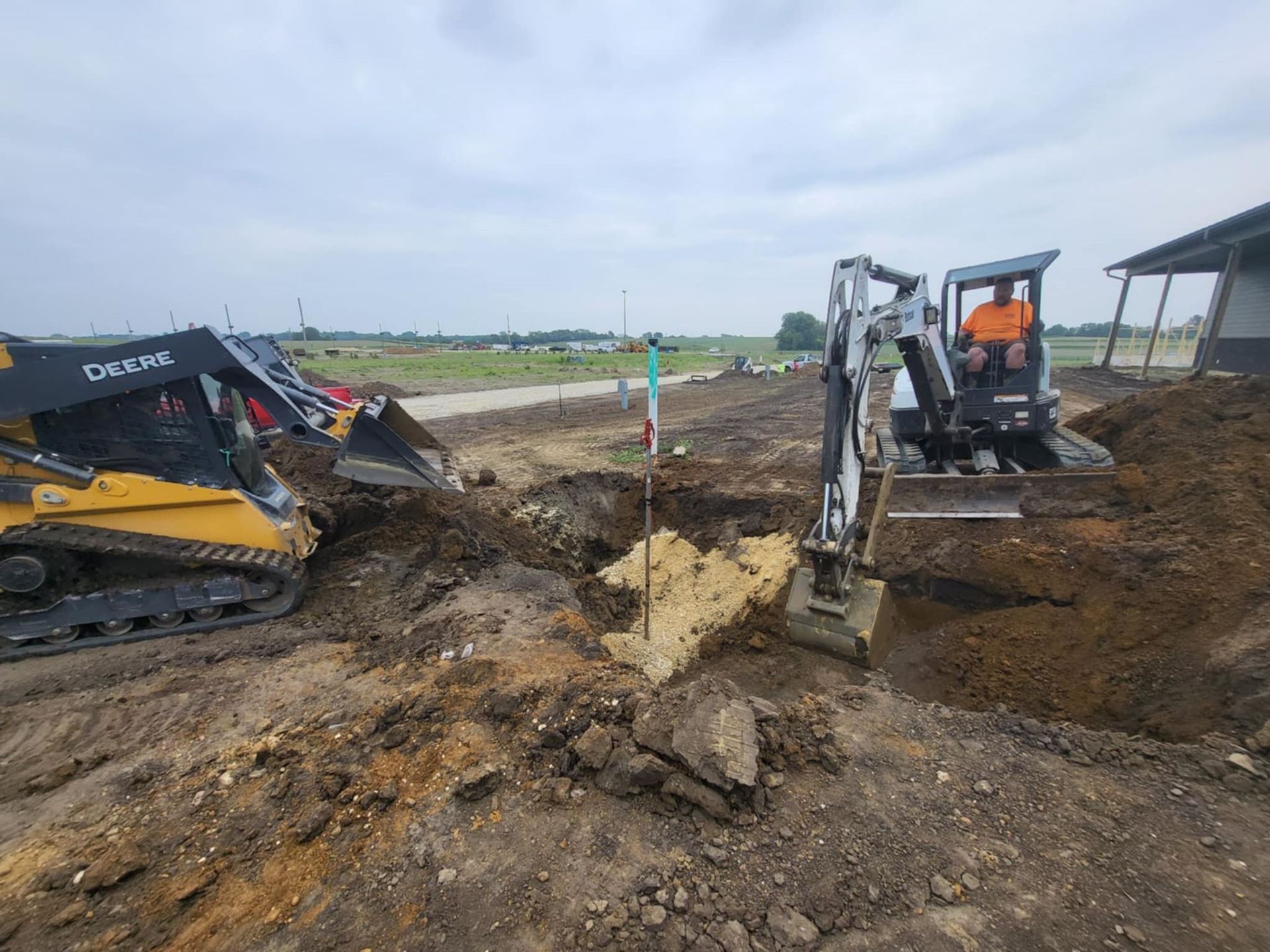 Two excavators digging a muddy trench at a roadside construction site under an overcast sky