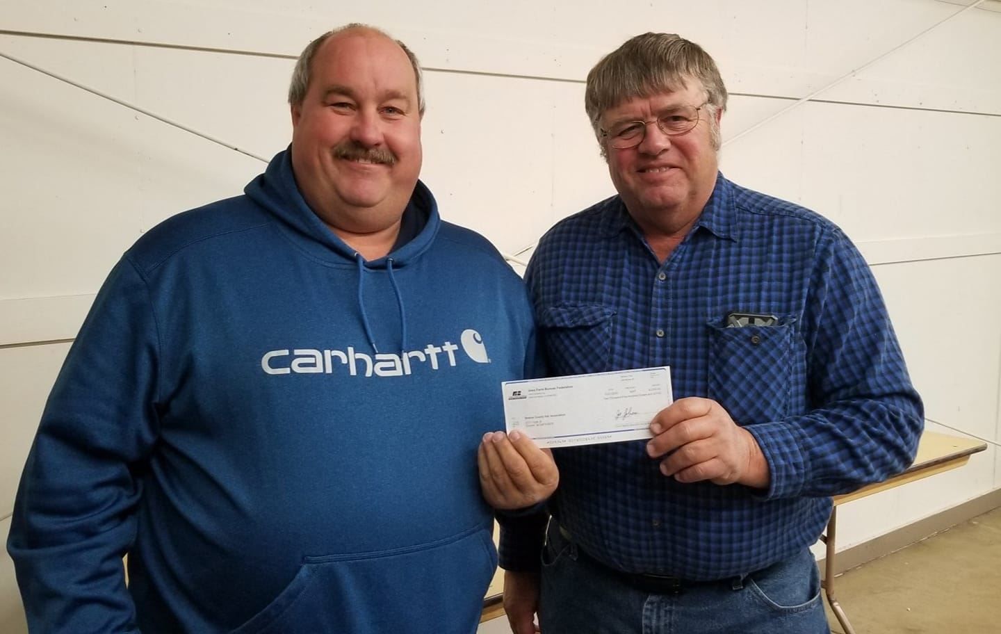 Two men indoors holding a check, one in a blue Carhartt sweatshirt and one in a blue checkered shirt.