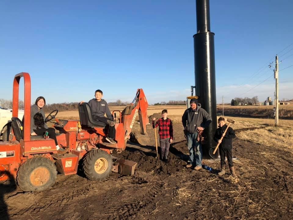 People with an orange excavator digging in muddy ground beside a tall black utility pole.