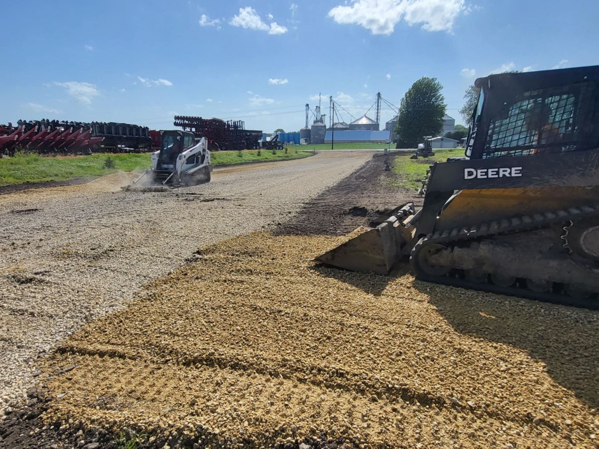 Construction equipment spreading gravel on a dirt road near industrial buildings on a sunny day