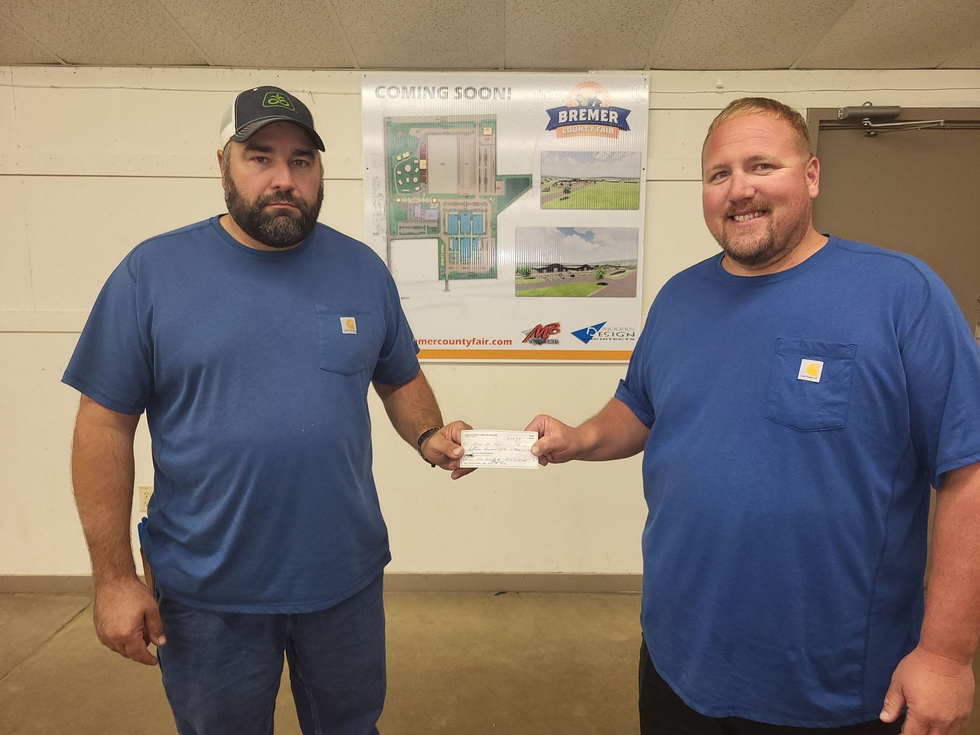 Two men in blue shirts stand indoors, holding a small check between them.