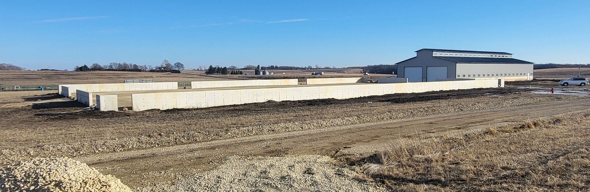 Wide dirt lot with long low white industrial buildings under a clear blue sky
