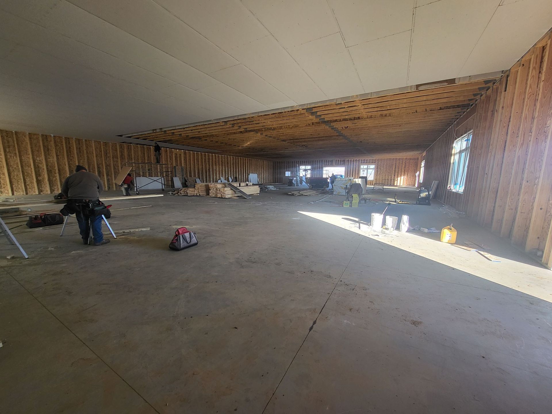 Large unfinished room with exposed wood framing, concrete floor, and a person working on the left.