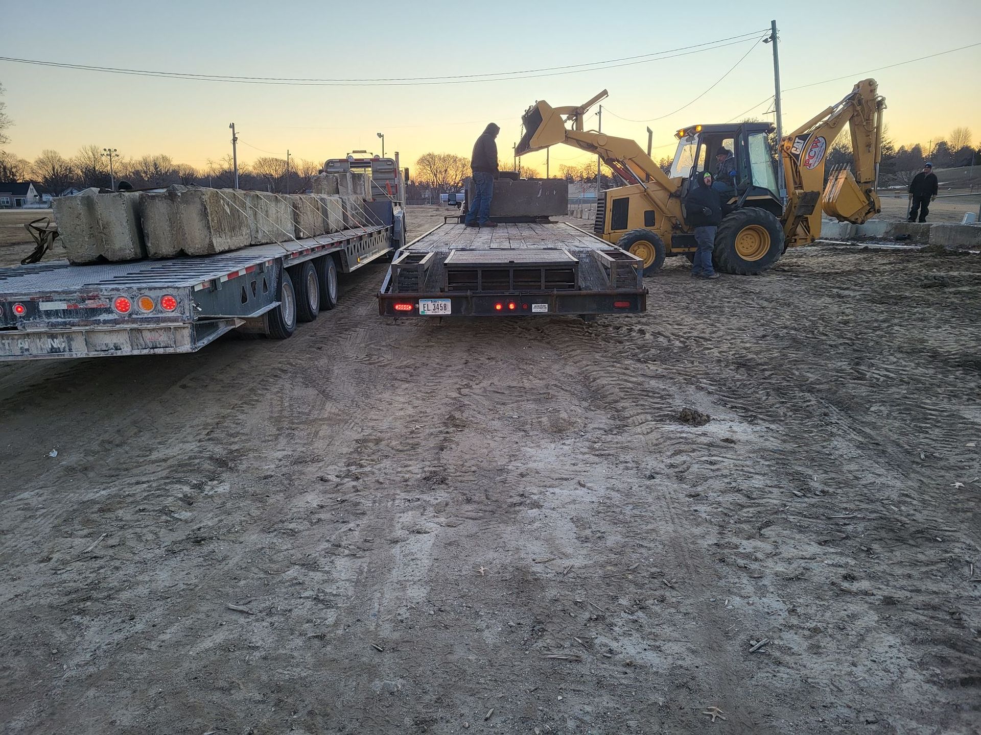 Construction site at dusk with a backhoe loading a flatbed trailer beside a truck.