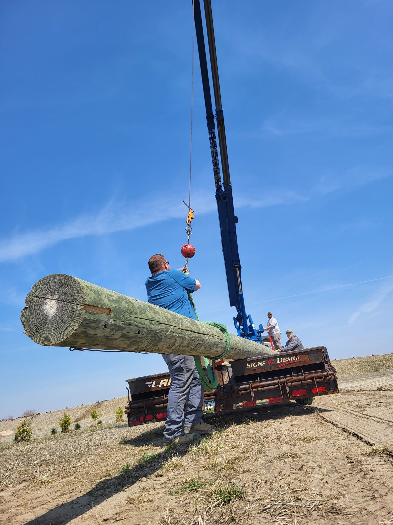 Worker guiding a large log being lifted by a crane onto a flatbed truck in a field