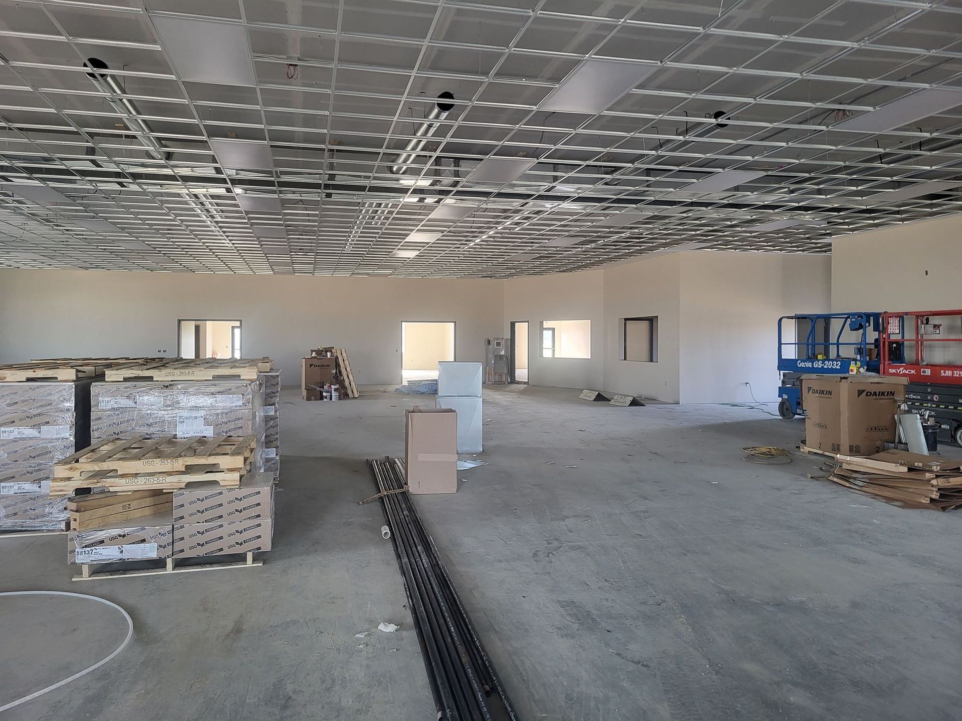 Empty commercial interior under construction with exposed ceiling grid, concrete floor, and stacked materials.